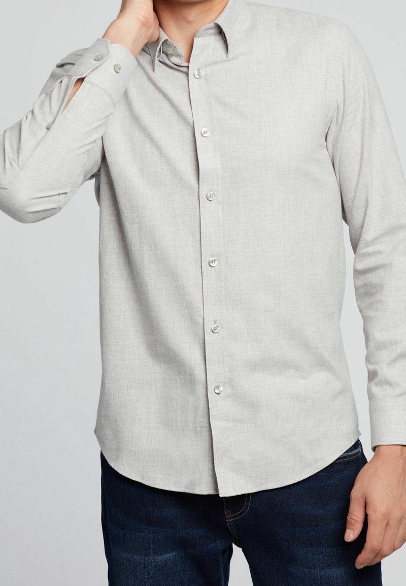 Homme portant une chemise gris clair à boutons et un jean foncé, main touchant le cou, sur fond uni.