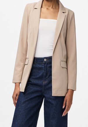 Mujer con blazer beige sobre top blanco sin tirantes y jeans azul oscuro de tiro alto, de pie sobre fondo blanco.
