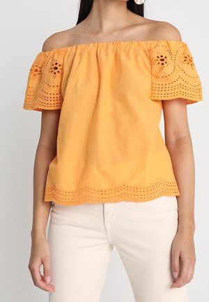 Kvinde iført en lys orange skulderfri top med broderede øjeldetaljer på ærmer og kant, kombineret med hvide bukser.