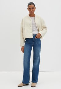 Veste courte en crème texturée avec un col rond et une fermeture à boutons, associée à un jeans droit bleu taille haute et des chaussures plates beiges.