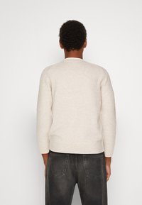 Suéter bege claro em tecido suave e texturizado, com gola redonda e barra canelada, combinado com jeans cinza escuro com bolsos traseiros.