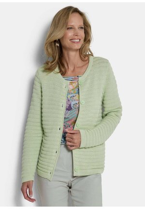 Vrouw glimlachend, gekleed in een lichtgroen gebreid vest over een kleurrijke bloemen top en lichtgrijze broek, staande tegen een witte achtergrond.