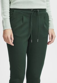 Dark green sweatpants with a drawstring closure, featuring pleats at the waist and soft, smooth fabric. Accents include metallic tips on drawstrings.