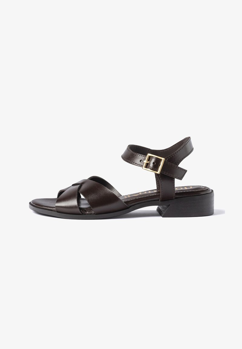 Sandale en cuir marron foncé avec un petit talon bloc, des brides croisées à l’avant et une bride de cheville avec une fermeture à boucle dorée.