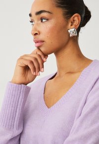 Pull en V lavande avec des poignets côtelés, texture douce, associé à une boucle d'oreille carrée en argent brillant avec un motif en tourbillon.