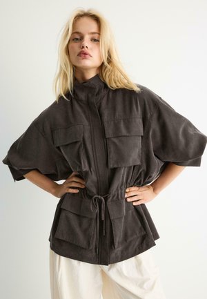 Blondynka w ciemnoszarej, oversize'owej kurtce z dużymi przednimi kieszeniami i białych spodniach, stoi z rękami na biodrach na białym tle.