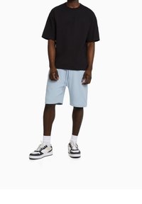 Muž v černém oversized tričku, světle modrých šortkách po kolena, bílých ponožkách a černo-bílých teniskách stojící na bílém pozadí.