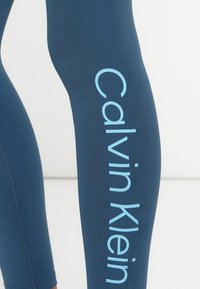 Leggings azuis feitos de um tecido elástico, com um proeminente logótipo "Calvin Klein" azul claro ao longo do lado e uma textura elegante e suave.