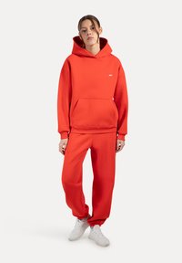 Felpa rossa con cappuccio e pantaloni della tuta coordinati realizzati in tessuto morbido. Caratterizzati da una tasca frontale, polsini a costine e dettaglio del logo bianco. Abbinati a sneakers bianche.