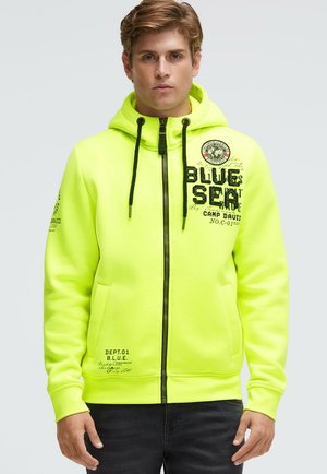 Neon-gelber Zip-up-Hoodie mit schwarzen Kordeln, Seitentaschen und bedruckten Designs auf Brust und Ärmel. Weicher, glatter Stoff.