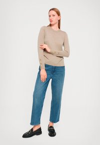 Beige Strickpullover mit langen Ärmeln und Knopfakzenten an den Bündchen, kombiniert mit lockeren blauen Jeans und schwarzen Slippern vor einem hellen Hintergrund.