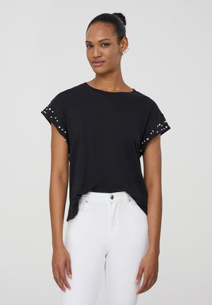T-shirt nera con maniche arrotolate, caratterizzata da borchie argentate lungo i bordi, abbinata a jeans bianchi. Materiale in cotone morbido, vestibilità comoda.