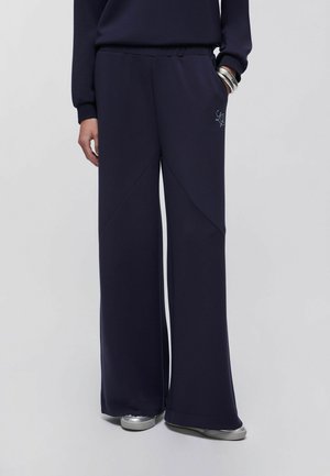 Femme portant un pantalon large bleu marine avec un logo brodé sur la poche, assorti à des baskets argentées et un haut noir à manches longues.