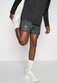 Black athletic shorts with a textured pattern and gold mountain graphic, paired with white sneakers and ribbed white socks.