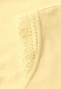 Detailní záběr na světle žlutou tkaninu s proužkovým lemem a vlnkovým krajkovým detailem, zdůrazňující texturu a švy.