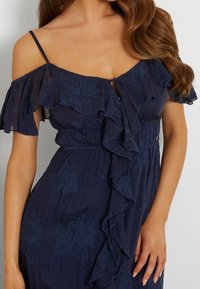 Navy blue dress featuring off-the-shoulder ruffles, embroidered floral patterns, and a keyhole detail at the front. Soft, textured fabric.