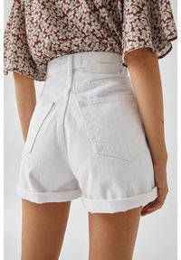 Femme portant un short en jean blanc avec ourlet roulé et un haut marron à manches courtes fleuri, vue de dos, de la taille au milieu des cuisses.