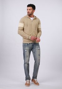 Beige zip-up hoodie met crèmekleurige accenten, gecombineerd met versleten blauwe jeans en bruine sneakers, tegen een witte achtergrond.