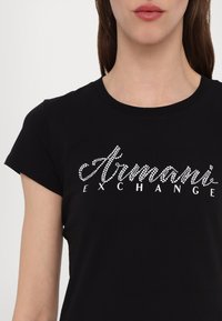 Schwarzes Baumwoll-T-Shirt mit Rundhalsausschnitt und kurzen Ärmeln, verziert mit Strassschriftzug "Armani Exchange" in Weiß.