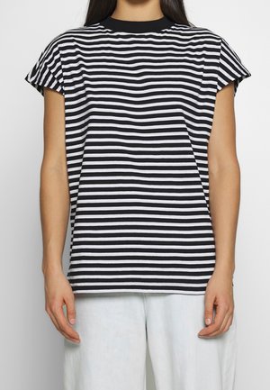 Persoon die een zwart-wit horizontaal gestreept T-shirt met korte mouwen draagt en een lichtblauwe broek, staande tegen een effen achtergrond.