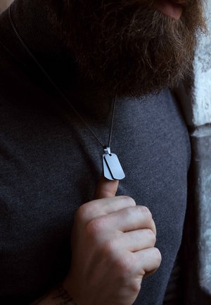 Zwart metalen dogtag ketting met twee gelaagde tags, voorzien van een matte afwerking en een dunne ketting, gedragen over een donkerkleurige coltrui.