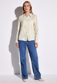Camicia in lino verde pallido leggermente texturizzata, con chiusura a bottoni e due tasche sul petto, abbinata a jeans larghi blu e ballerine crema.