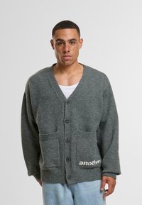 Siva volnena cardigan s V-izrezom, šestimi gumbi, dvema sprednjima žepoma in malim logotipom na robu. Tkanina ima mehak teksturo.