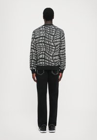 Črna in bela sweatshirt z drznim, ponavljajočim se tekstovnim vzorcem, z okroglim izrezom in rebrastimi manšetami, v kombinaciji s črnimi hlačami in supergami.