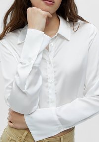 Chemise blanche à manches longues avec un col classique, confectionnée dans un tissu lisse, avec des détails à boutons et des poignets larges.