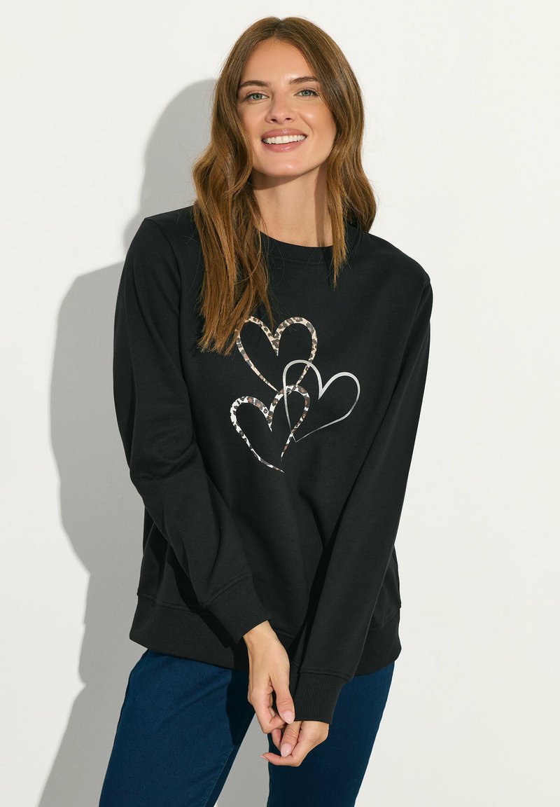 Sudadera negra con cuello redondo y mangas largas, que presenta tres diseños de corazones en acentos metálicos. Combinada con pantalones azul oscuro.
