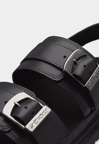 Μαύροι λουράκια δερμάτινων σανδαλιών με ασημένιες μεταλλικές αγκράφες χαραγμένες με την επιγραφή «Dr. Martens» σε κάθε αγκράφα.