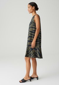 Sleeveless olive green dress with dark blue abstract patterns, flowy fabric, knee-length with a flared hem. Worn with black heeled sandals.