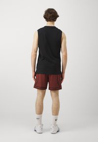 Camiseta sin mangas negra combinada con pantalones cortos rojos con acentos negros. El modelo lleva calcetas blancas y zapatillas. Fondo liso.
