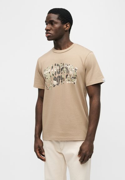 Άνδρας που φοράει μπεζ t-shirt Billionaire Boys Club με λογότυπο παραλλαγής, συνδυασμένο με ανοιχτόχρωμο παντελόνι, στέκεται μπροστά σε απλό φόντο.