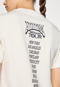 Kremo spalvos marškinėliai su juodu grafinio dizaino spaudiniu ant nugaros, kuriame užrašyta "Adidas Still Swinging Tour" ir miestų pavadinimai išdėstyti vertikaliai.