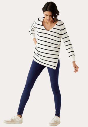 Túnica de punto a rayas en blanco y azul marino, cuello en V, mangas largas, dobladillo asimétrico. Combinada con jeans estrechos azul marino y zapatillas blancas.