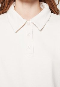 Camisa polo blanca con un cuello clásico, cuatro botones y una textura de tela suave. El cuello y los botones son detalles destacados.