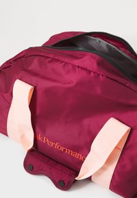 Dieprode nylon sporttas met een rits, voorzien van lichtroze handvatten en een geprint logo in oranje. Gladde textuur met een gestructureerde vorm.