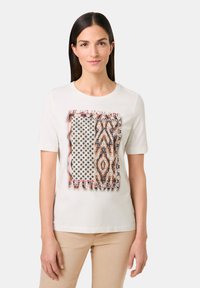 White cotton t-shirt featuring a printed design with various patterns: dots, stripes, and a decorative border. Short sleeves and crew neck.