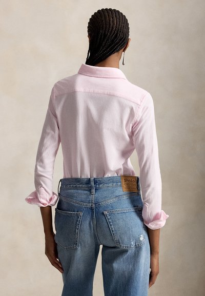 Persona con i capelli intrecciati che indossa una maglia a maniche lunghe rosa chiaro e jeans di denim blu, di spalle alla fotocamera su uno sfondo neutro.