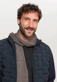 Giacca trapuntata blu navy, sciarpa grigia, camicia scura con bottoni. Sciarpa strutturata avvolta in modo rilassato, uomo sorridente con capelli ricci e barba.