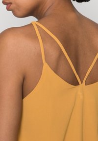 Top giallo senza maniche con spalline sottili e design a drappeggio sul retro, caratterizzato da una leggera lucentezza e una texture morbida.