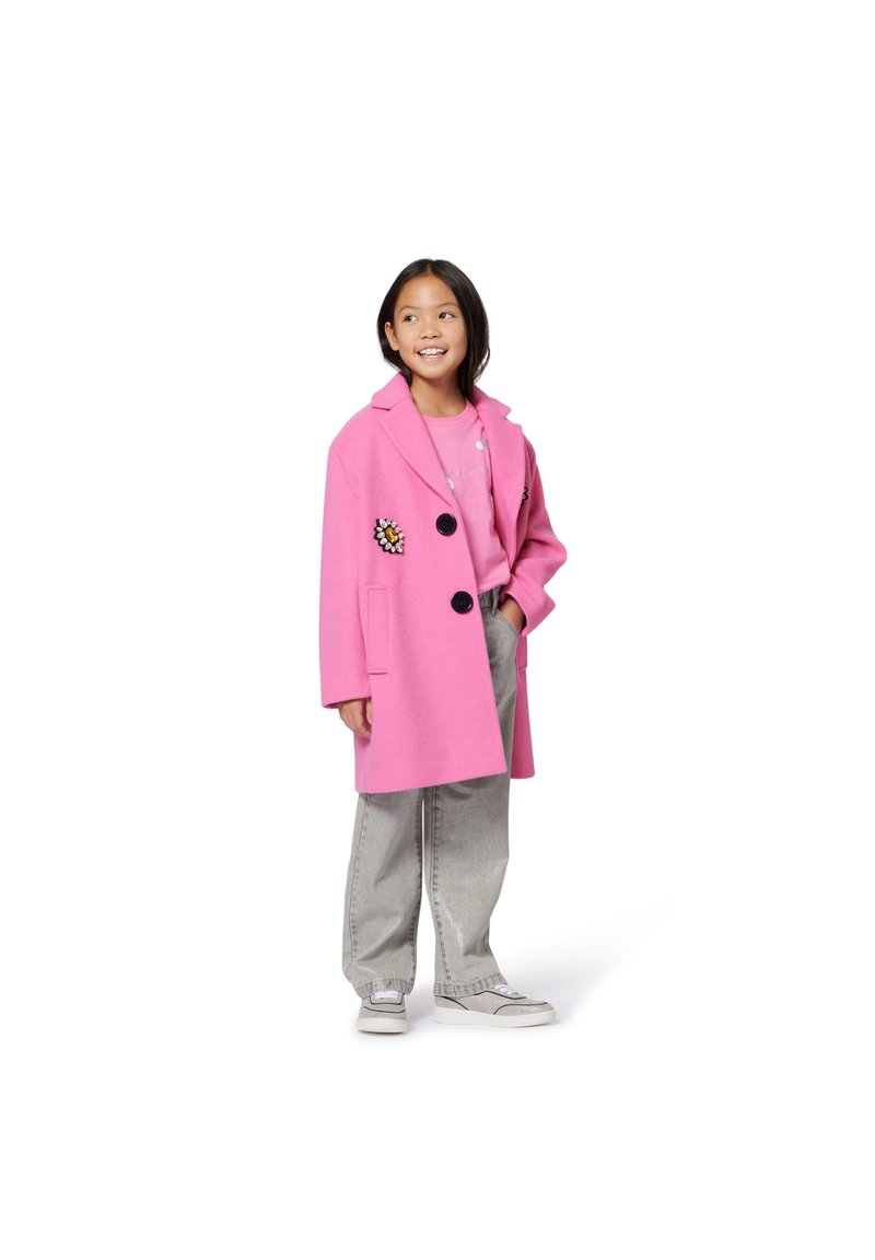 Rosa ullkappa med svarta knappar och blommig brodyr, kombinerad med en ljusrosa skjorta och grå vida jeans, fulländad med vita sneakers.