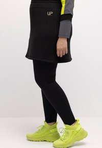 Persoon in zwarte leggings, een zwarte sportrok, een grijs en geel langemouwenshirt en felgele chunky sneakers, staande in zijaanzicht.