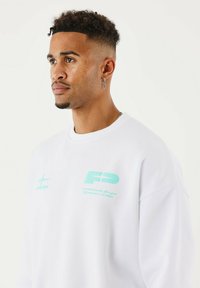 Hombre con cabello corto y rizado y perforaciones en la cara, vistiendo un sudadera blanca con logotipos en azul claro, mirando hacia la izquierda frente a un fondo claro y liso.