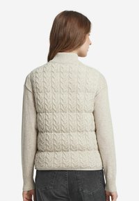 Frau, die einen beigen, ärmellosen Pullover mit Zopfmuster über einem langärmligen Pullover trägt, kombiniert mit dunkelgrauen Jeans, mit dem Rücken zur Kamera.