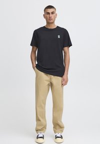 T-shirt noir à manches courtes avec un petit logo vert "S", associé à un pantalon beige et des baskets noir et blanc sur un fond neutre.