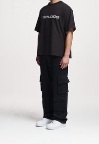 T-shirt noire oversize avec inscription blanche "STUDIOS", associée à un pantalon cargo noir avec plusieurs poches et des baskets blanches.