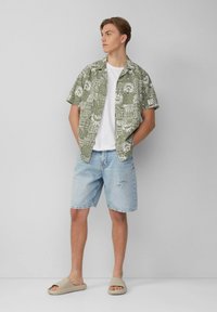 Korte mouwen groene patroon blouse met witte motieven, witte t-shirt, lichte spijkerbroek met een scheur en beige slipper sandalen.