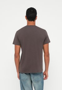 Mężczyzna odwrócony plecami, ubrany w gładką, ciemnoszaro koszulkę z krótkim rękawem oraz jasnoniebieskie dżinsy na tle czystego białego tła.